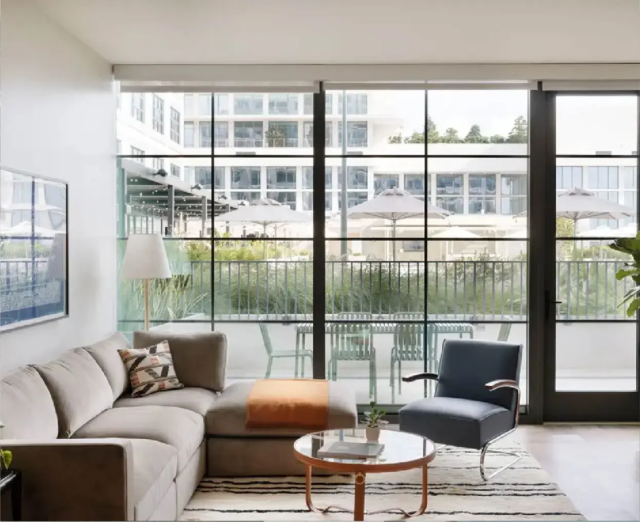 Living room with a beige sectional sofa, a black armchair, a round wooden coffee table, and a balcony with outdoor chairs and umbrellas.