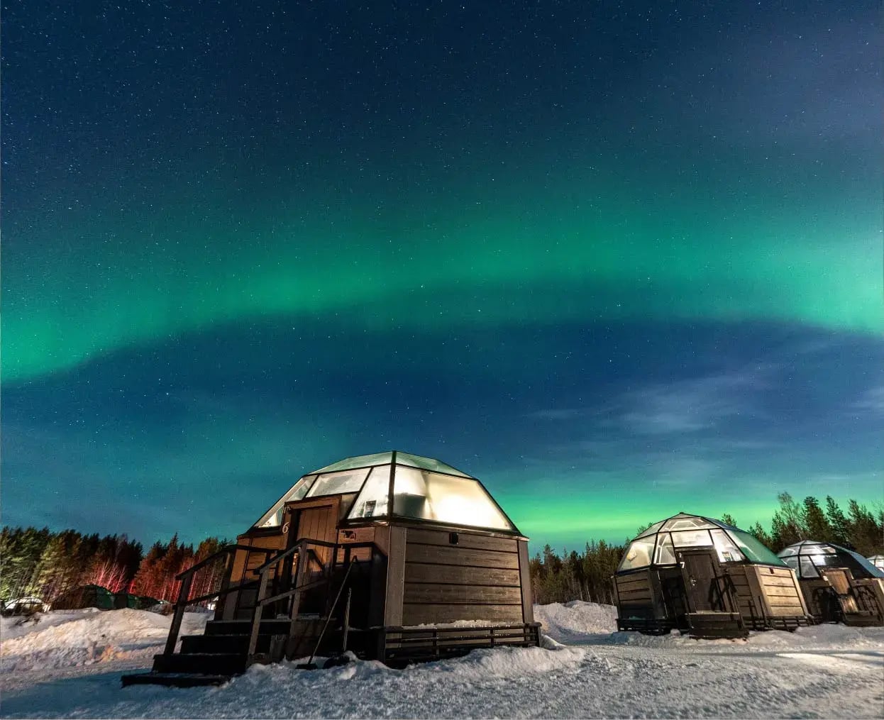 Gläserne Hütten unter den Nordlichtern in einer verschneiten Landschaft bei Nacht.