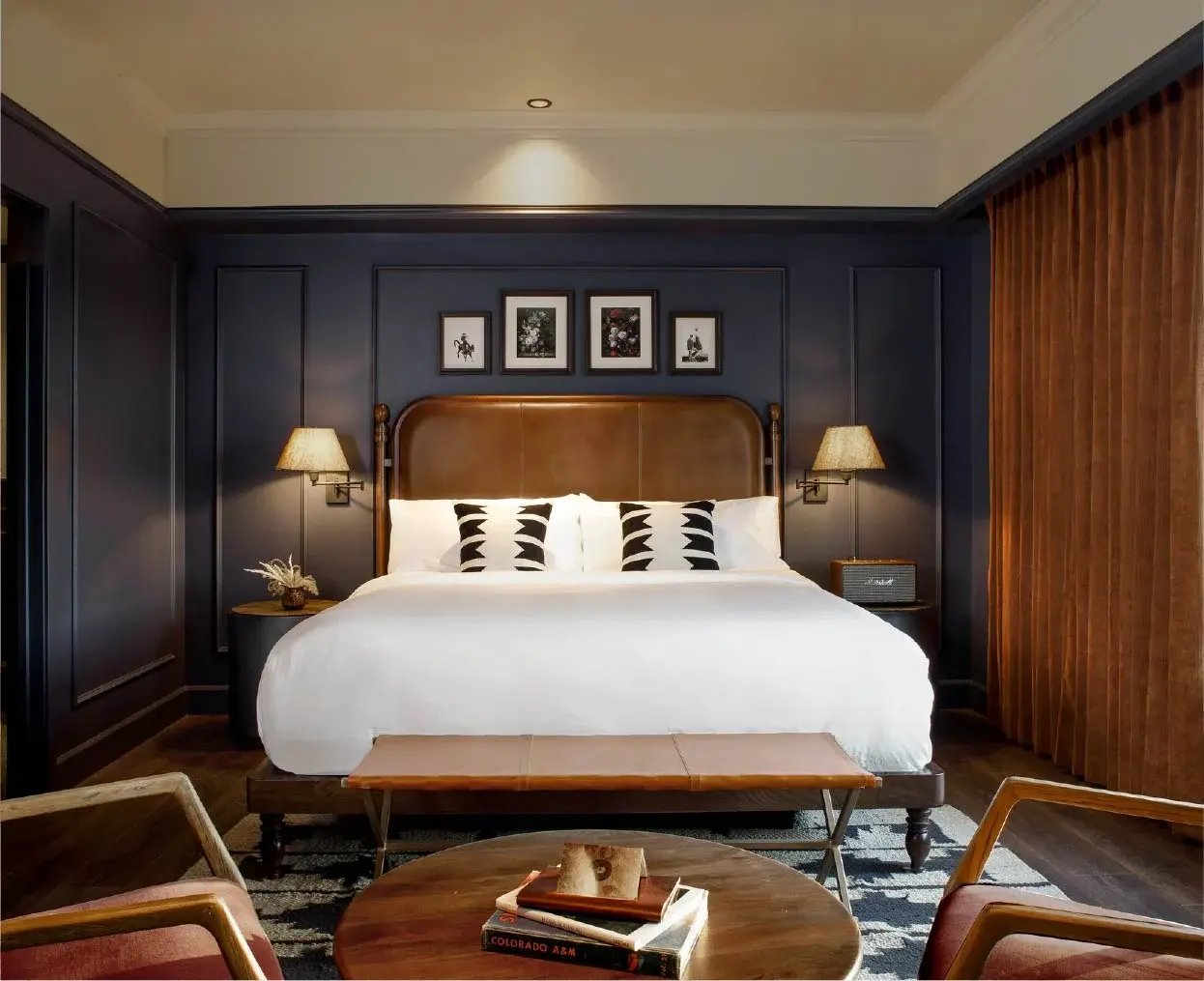 Ein Kingsize-Bett mit weißer Bettwäsche, schwarzen und weißen Kissen, dunklen Holzmöbeln und gerahmten Kunstwerken an einer marineblauen Wand in einem Schlafzimmer.