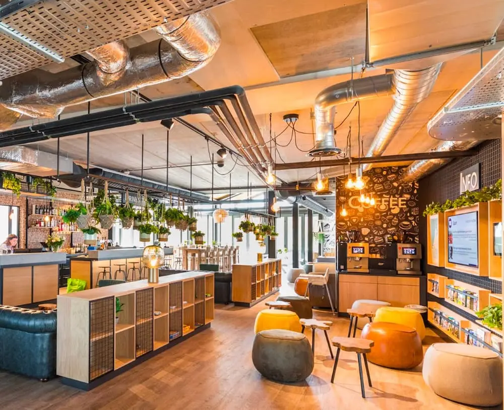 Moderno vestíbulo de hotel con plantas colgantes, zonas para sentarse, detalles de madera y conductos a la vista.