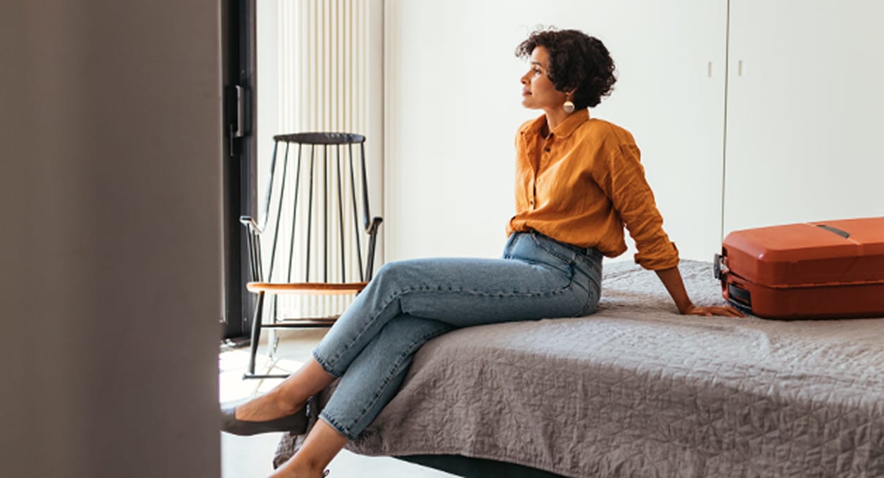 Woman sitting on a bed with a suitcase beside her, looking thoughtfully at a window or wall.