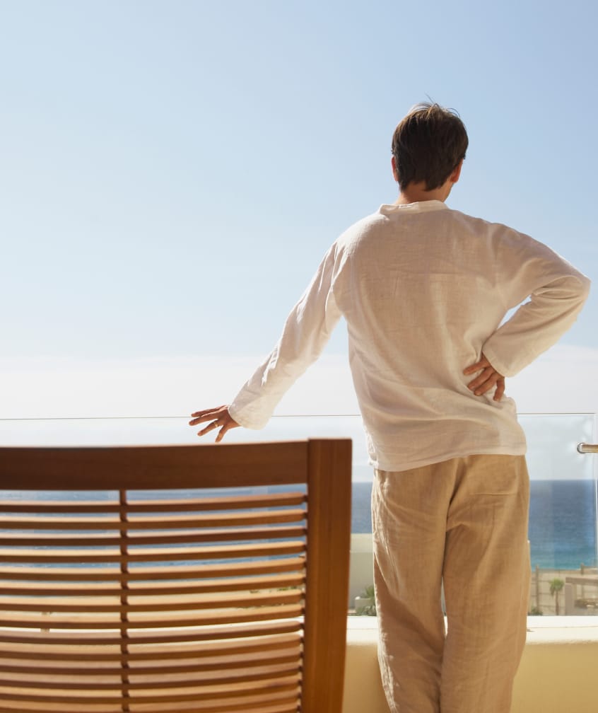 Eine Person, die auf einem Balkon steht und auf das Meer schaut, mit einer Hand am Geländer und der anderen an der Hüfte.
