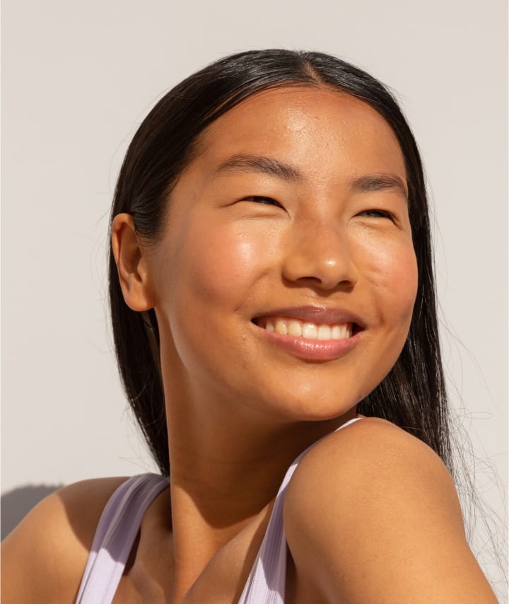 Smiling woman with long black hair, wearing a white tank top, turned slightly to the side against a plain background.