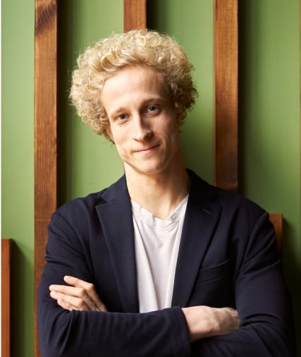 Junger Mann mit lockigen blonden Haaren in einem dunklen Blazer, der seine Arme verschränkt, steht vor einem grünen Hintergrund mit vertikalen Holzplatten.