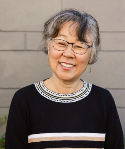 Smiling elderly woman with short, gray hair and glasses, wearing a black sweater with white detailing, standing against a gray wall.