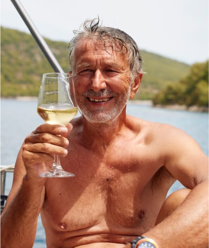 Ein lächelnder älterer Mann mit grauen Haaren und Bart, ohne Hemd, hält ein Glas Weißwein im Freien in der Nähe eines Gewässers.