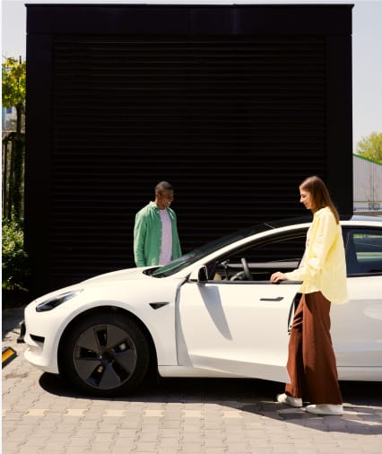 Zwei Frauen stehen neben einem weißen Elektroauto, das vor einem schwarzen Garagentor geparkt ist.