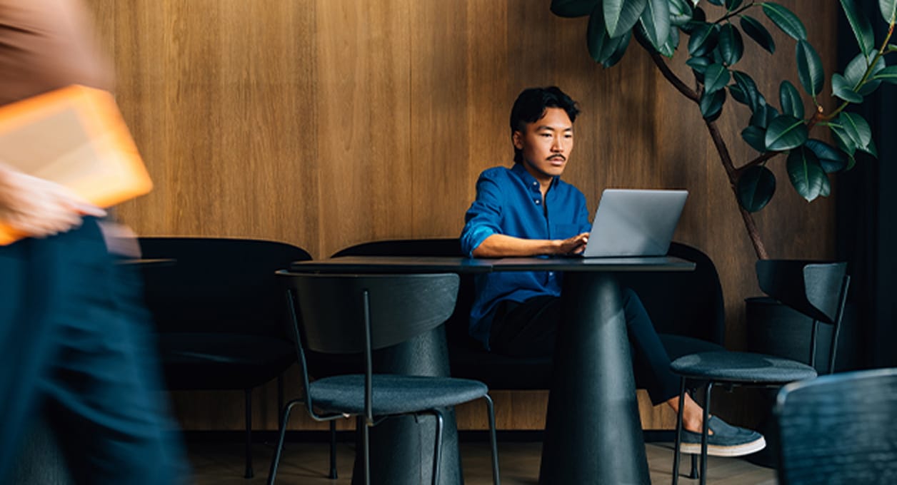 Ein Mann in einem blauen Hemd arbeitet an einem Laptop an einem schwarzen Tisch in einer modernen, gemütlichen Einstellung mit Holzwänden und Grünpflanzen.