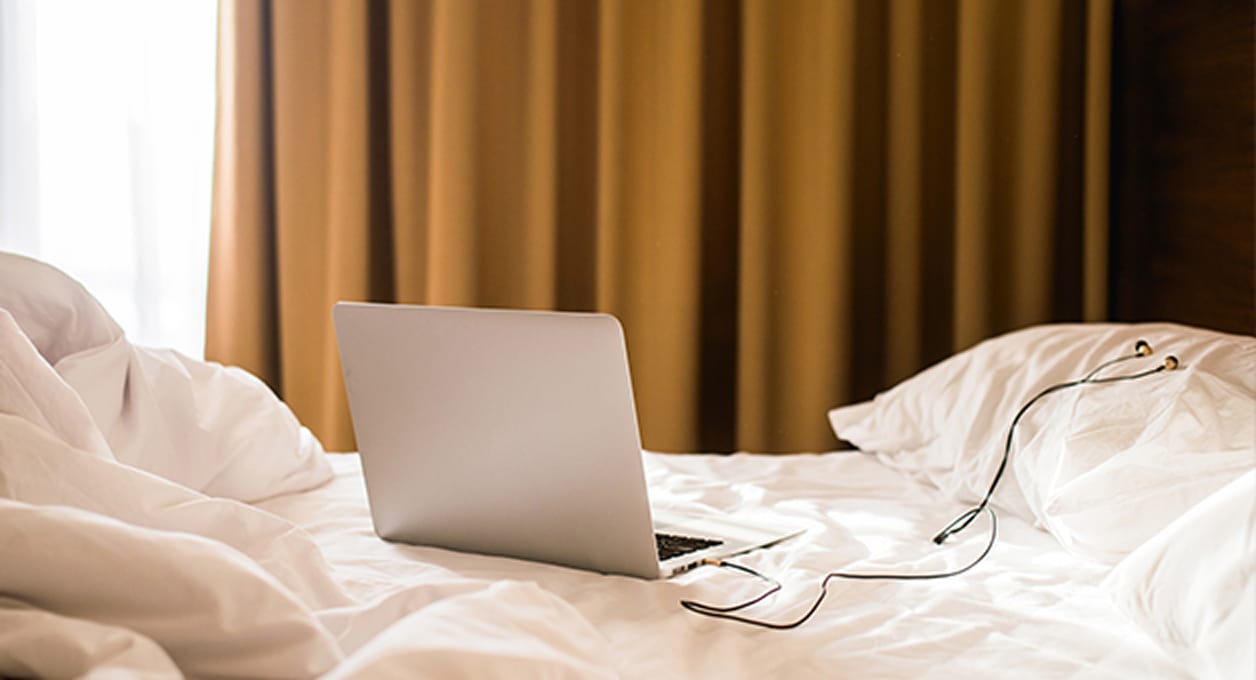 Laptop with earphones resting on rumpled white bedsheets in a bedroom.