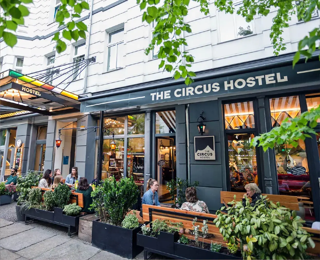 Des personnes assises et discutant à l'extérieur de l'auberge Circus, dans une rue de la ville, avec un feuillage vert au-dessus de la tête.
