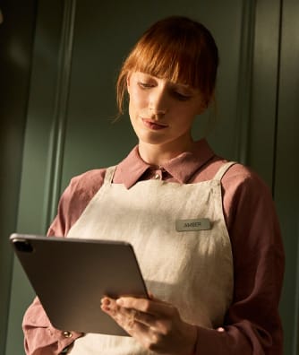 A woman in a pink blouse and beige apron looks down at a tablet, standing in front of green paneled walls.